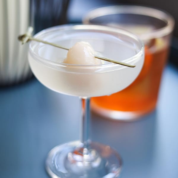 Lychee martini in a coupe glass with a lychee garnish, next to an orange drink.