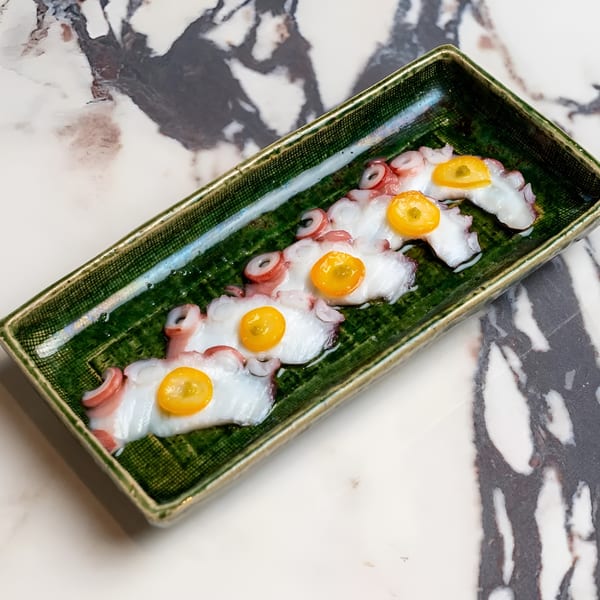 Thinly sliced octopus with yellow kumquat on a green rectangular plate, on a marble surface.