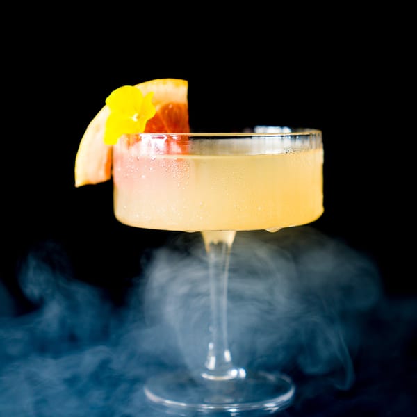 A vibrant cocktail in a coupe glass, garnished with grapefruit and a yellow flower, surrounded by mist.