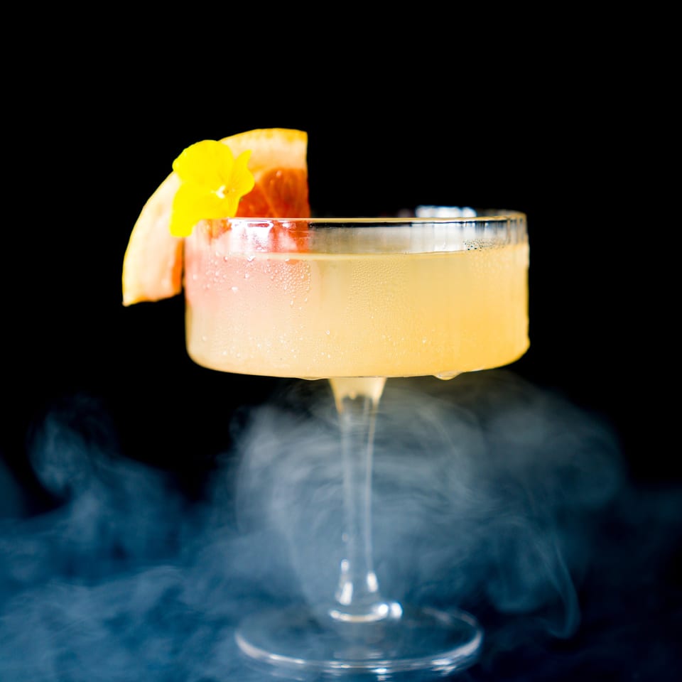 A vibrant cocktail in a coupe glass, garnished with grapefruit and a yellow flower, surrounded by mist.