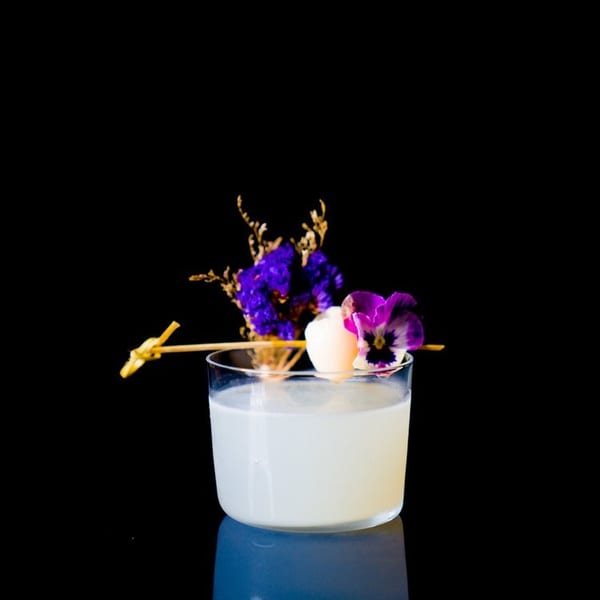 A light-colored cocktail garnished with purple flowers and a lychee on a dark background.