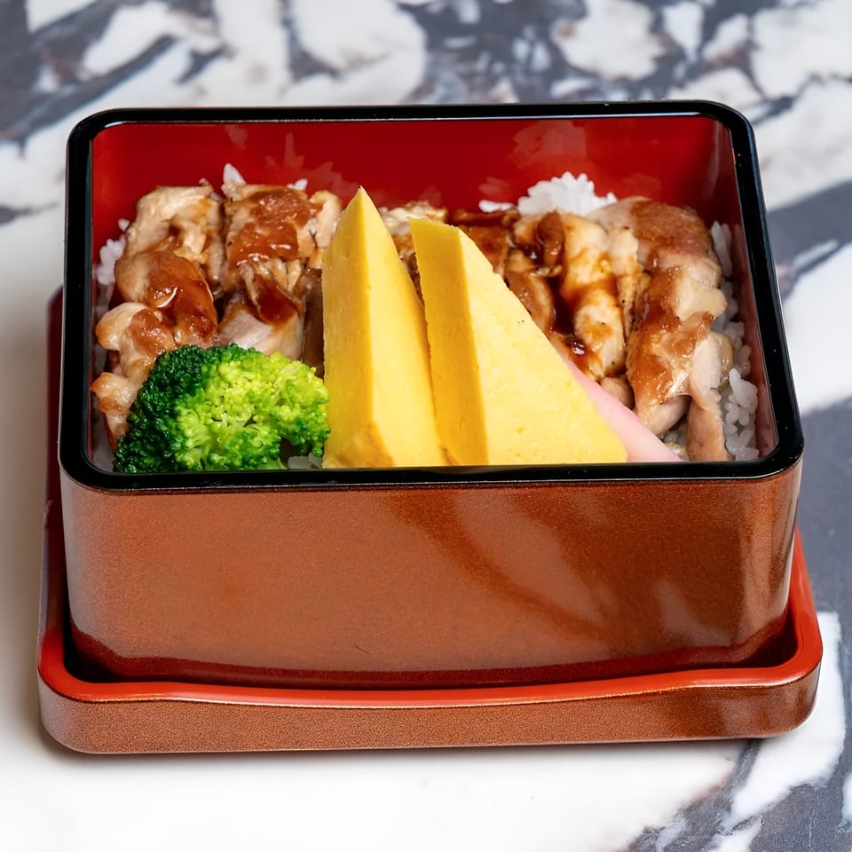 A bento box filled with teriyaki chicken, white rice, broccoli, and two pieces of tamagoyaki.