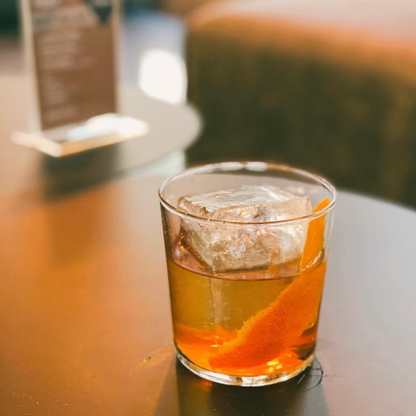 An Old Fashioned cocktail with a large ice cube and orange peel on a dark table.
