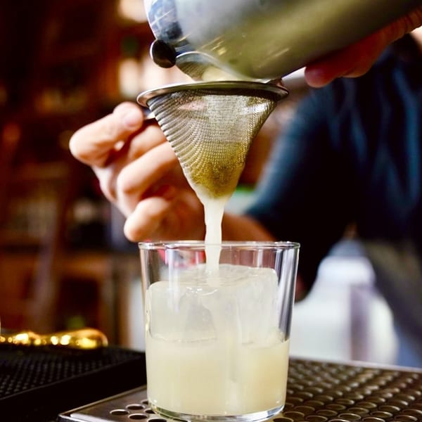 Bartender straining a light-colored cocktail from a shaker into a glass with ice.